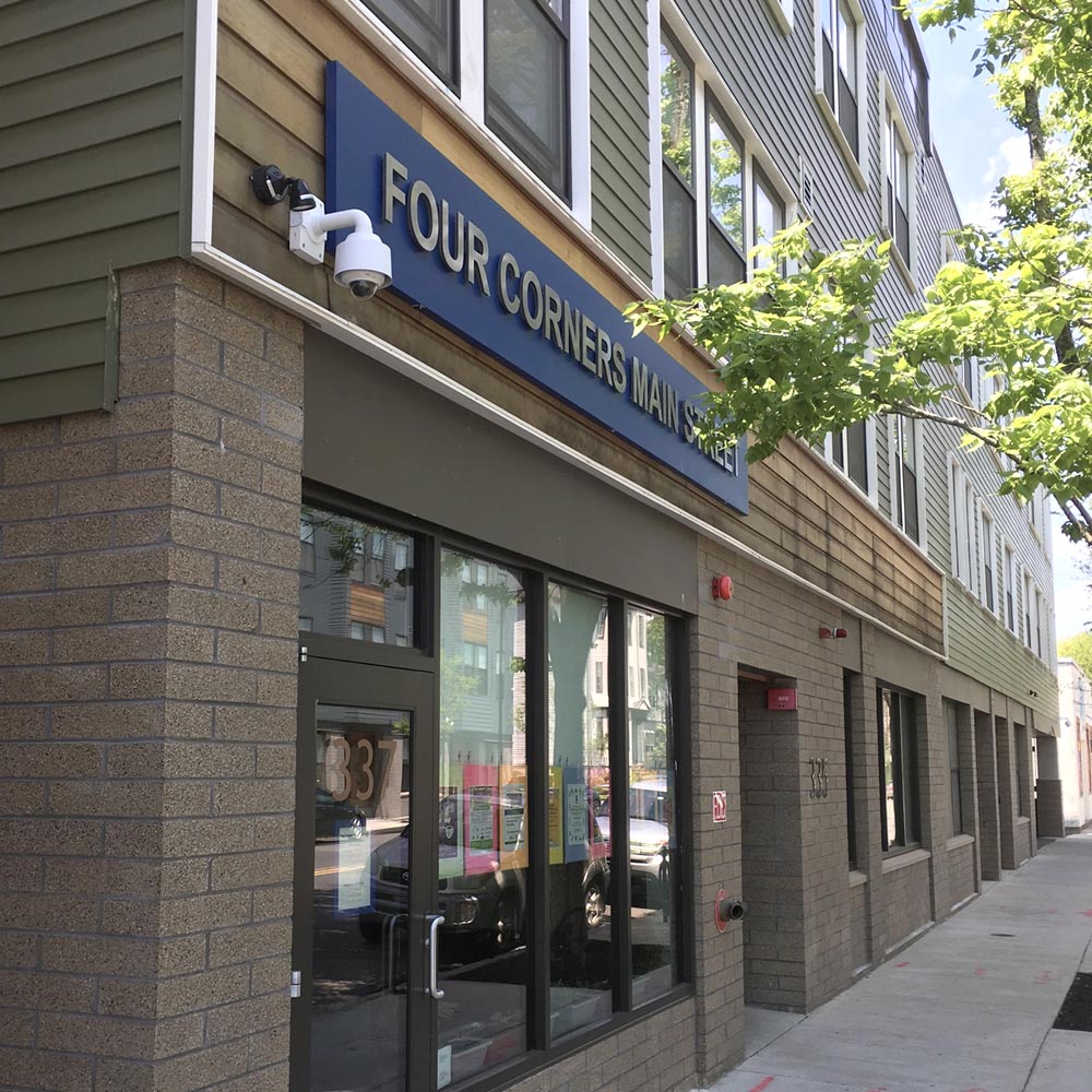 Sidewalk and building exterior with windows and bring of Four Corners Main Street Office with tree in corner touching building